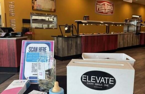 A reception area with a "Pizza Ranch" sign on the wall, a counter with trays, and a table with a water bottle, a white box labeled "ELEVATE," brochures, and a QR code sign.