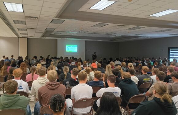 Audience seated in a conference hall facing a projector screen with a speaker standing at the front under flat ceiling tiles.