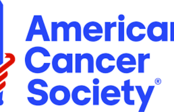 A person in a green uniform with a yellow hat is holding a large check, standing outdoors next to a man in a beige uniform with a red tie.