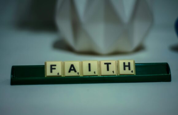 Scrabble tiles spelling out "FAITH" on a green rack, with blurred background including a white geometric object and part of a Pizza Ranch uniform.
