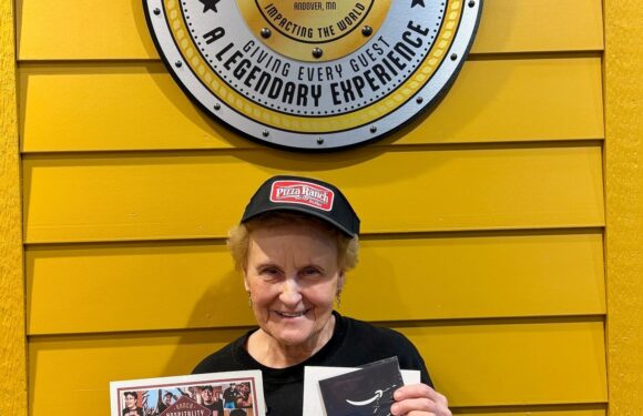 Smiling person in a black Pizza Ranch cap holds a congratulatory card and photo while standing against a yellow wall with a Pizza Ranch sign.