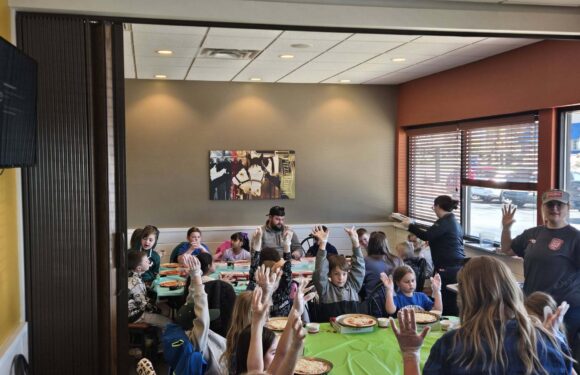 Children gather around a table with pizza while adults stand nearby, hands raised in a casual indoor birthday party setting