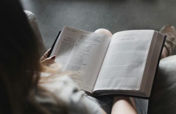 A person reads a large open book while sitting on a couch, with a focus on the book's pages and their hand holding the edge.