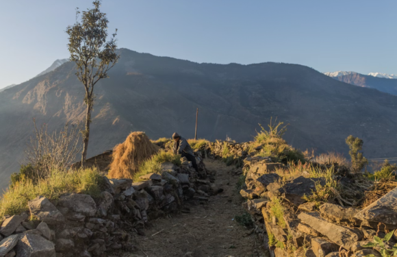 A person in a hoodie and dark pants climbs a rocky, mountain trail with a landscape of mountains, some snow-capped, in the background during sunset.