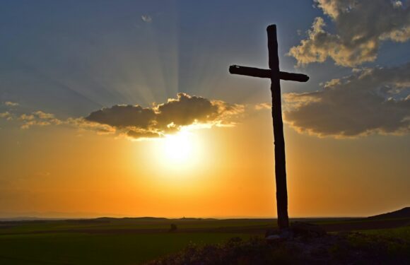 Dark cross silhouette on a hillside at sunset, glowing clouds and golden sky with sun rays behind it.