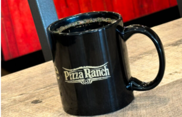 A black coffee mug with the Pizza Ranch logo on it, filled with dark coffee, placed on a wooden table with a red and black background.