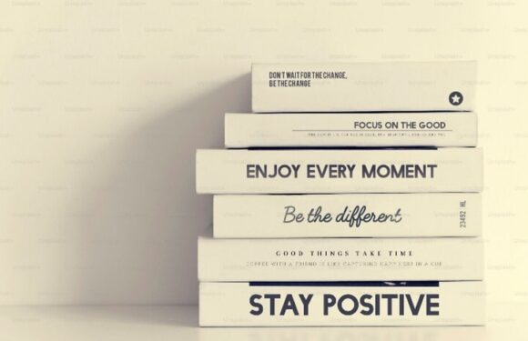 Stack of motivational books on a table with titles “ENJOY EVERY MOMENT,” “Be the different,” and “STAY POSITIVE,” plus “FOCUS ON THE GOOD.”