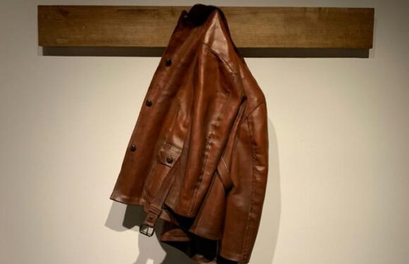 Brown leather jacket hanging on a wall mount beam against a plain light background.