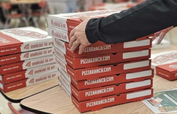 Stacked pizza boxes on a table with a person reaching for one, in a fast food restaurant with red chairs and people in the background.