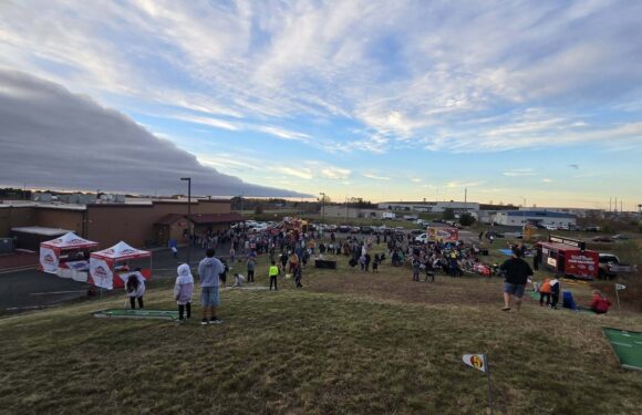 Crowd gathers on a grassy field at an outdoor fun event near buildings, with food booths and trucks under a sunset sky.