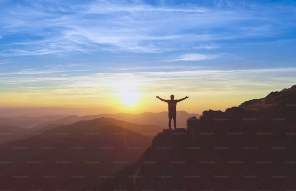 A person standing on a mountain peak at sunrise or sunset with arms outstretched, overlooking distant mountain ranges under a partly cloudy sky.