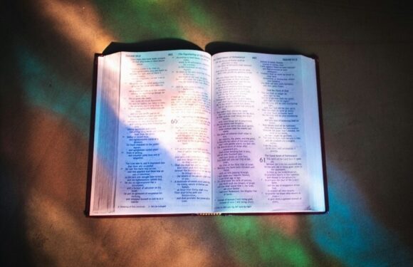 An open Bible on a surface, illuminated by colorful light reflections, with visible text and a dark background.