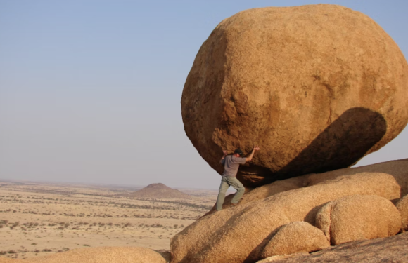 A person in a gray shirt and pants pushes a large boulder on a rocky desert landscape with sparse vegetation and distant low hills.