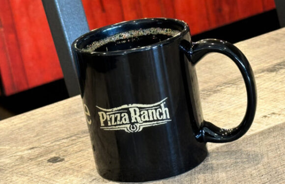 A black ceramic mug filled with a dark beverage, featuring the Pizza Ranch logo, sits on a wooden table with a wood-paneled red wall in the background.