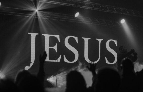 A black-and-white photo of a concert or event with the word "JESUS" displayed on a screen or backdrop. Audience members are visible with raised hands.