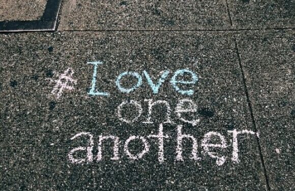Chalk message on pavement: “Love one another” written in blue and white letters on dark asphalt.