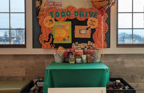 A food drive setup with a basketball-themed backdrop, baskets of food on a green-covered table, and a large WAFER food pantry sign in front. Two inflatable basketballs hang above.