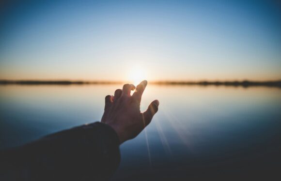 Silhouette of a person’s hand forming a heart shape at calm lakeside during sunrise, warm light reflecting on the water.