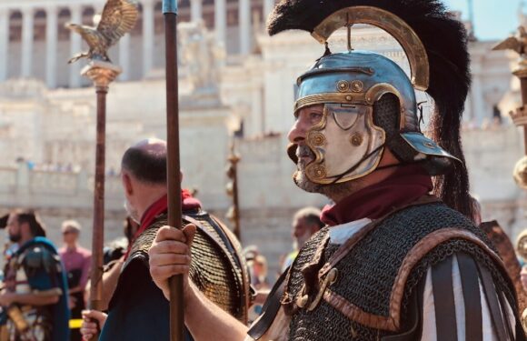 Reenactor in ornate Roman-style armor and helmet with blue crest holds a spear in a sunny ancient-stadium setting.