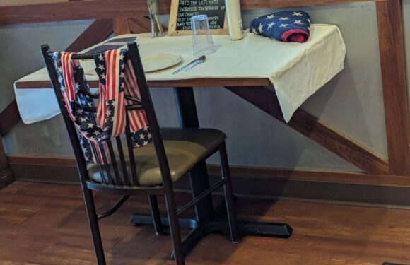 Dining setup with small table covered in white cloth, American flags draped over chairs, bunting and a “fallen comrade” sign by a window.