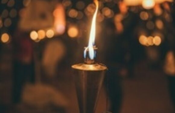 A lit candle in a holder with a flame, blurred warm lights and silhouettes of people in the background.