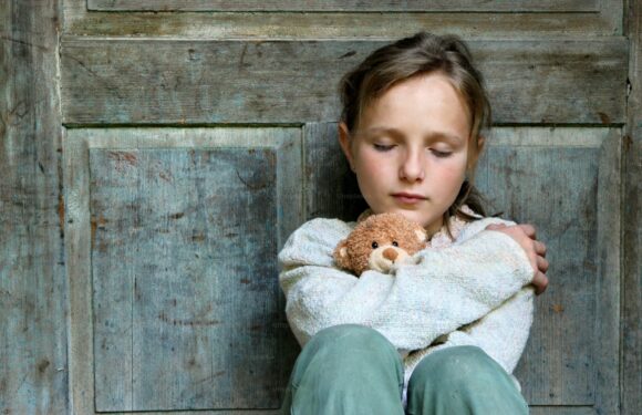 A young girl with closed eyes sitting against a rustic wooden background, hugging a small teddy bear with a neutral expression.