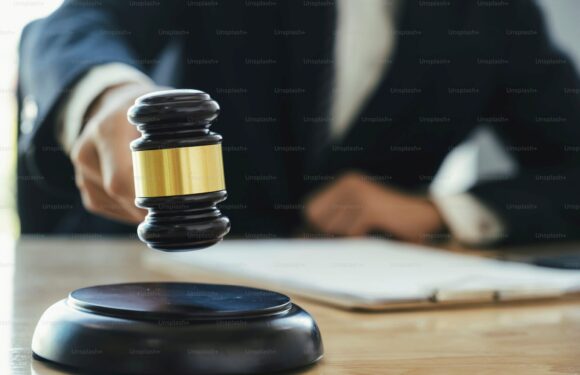 A person in a suit strikes a gavel on a table, with a clipboard in the background, symbolizing a legal or judicial setting.