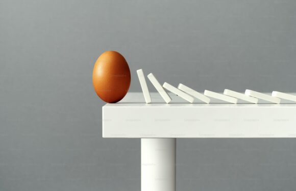 A brown egg topples as white dominoes fall off a white table against a neutral background.