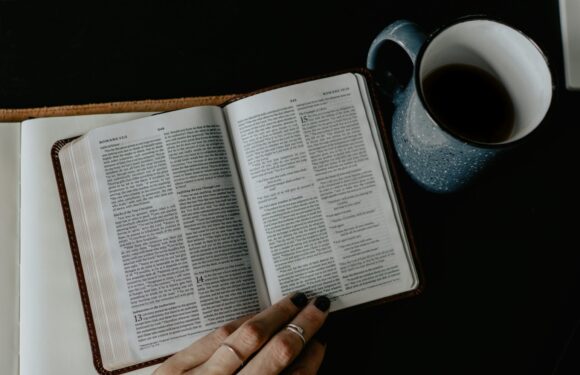 An open Bible with a hand turning the pages, placed on a dark surface, next to a blue ceramic mug filled with black coffee.