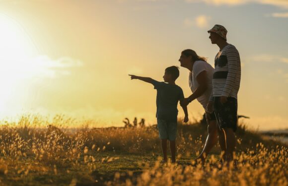 A family of three walking on a grassy path during sunset, with the child pointing ahead, in a field with tall grass and a partly cloudy sky.