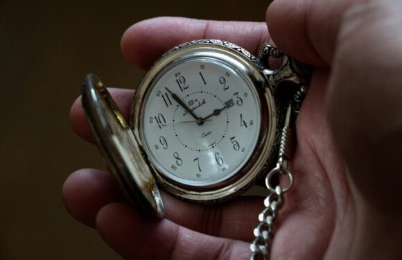Close-up of a person's hand holding an ornate pocket watch with a white face, black numbers, and a chain, showing the time as 2:12.
