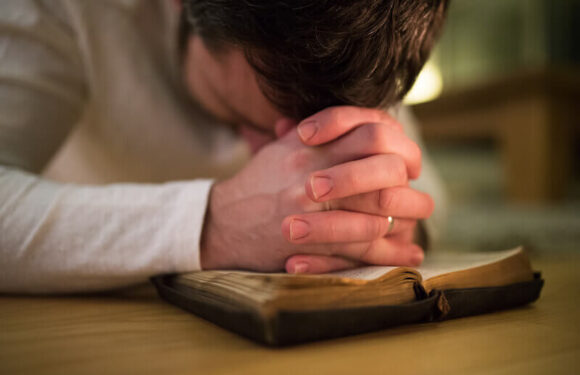 Person praying or reflecting with hands clasped over an open Bible on a wooden surface, in a warmly lit indoor setting.