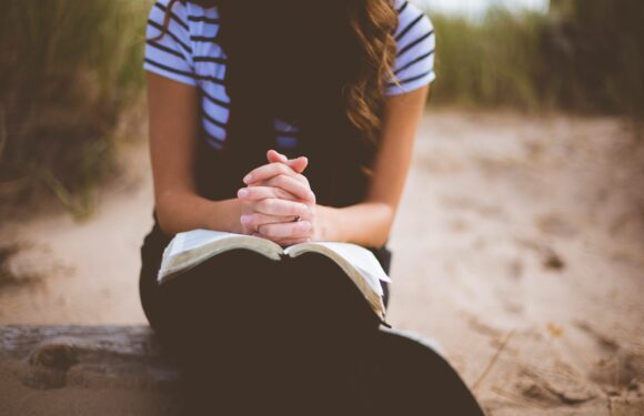 A woman with long hair, wearing a striped shirt, sits outdoors with her hands clasped in prayer over an open Bible on her lap, on a sandy surface with grass in the background.