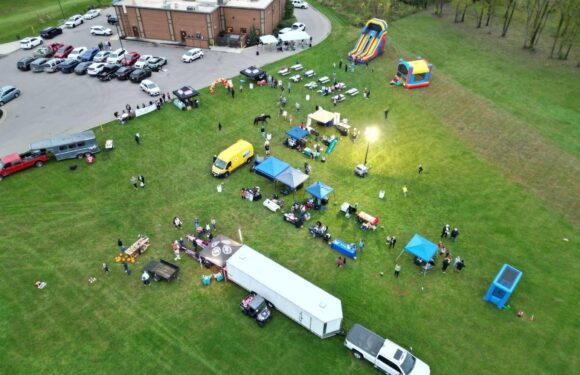 An outdoor gathering with tents, tables, and inflatable slides on a grassy field near a parking lot and a brick building, with people and vehicles present.