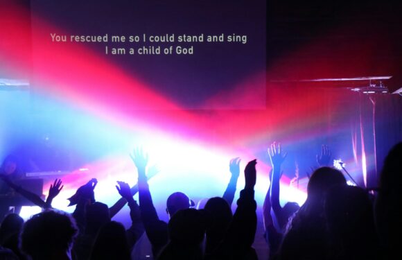 People with raised hands are gathered in front of a large screen displaying the lyrics: "You rescued me so I could stand and sing I am a child of God," in a dark venue with colorful lighting.