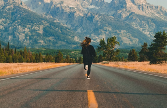 A person walking on an empty road surrounded by fields with mountains in the background during daytime.