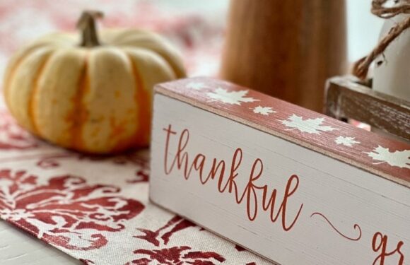 A white and pink wooden sign with the word "thankful" written in cursive, placed on a red and white patterned tablecloth, with a small pumpkin in the background.