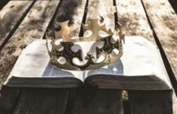 A silver crown with ornate cut-out designs rests on an open book on a wooden table with a white cloth underneath.