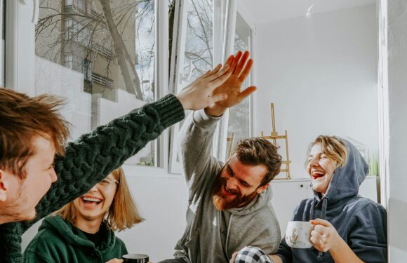 Four people are sitting together in a cozy room, smiling and enjoying coffee, with two of them giving a high-five. There's a teapot and snacks on the table.