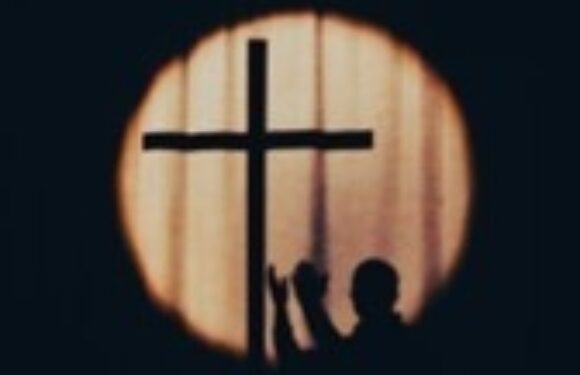 Silhouette of two people kneeling with raised hands in prayer, viewed through a circular cross-shaped window with a wooden background.