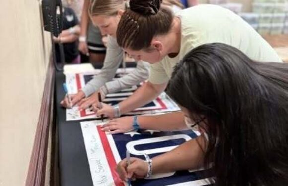 People gathered in a school room writing and signing large patriotic banner flags and posters, with a group leaning over tables.