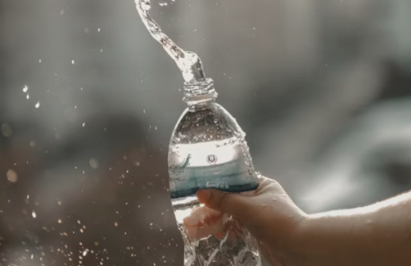 A hand squeezing a clear plastic water bottle causing water to spray out, with blurred urban background.