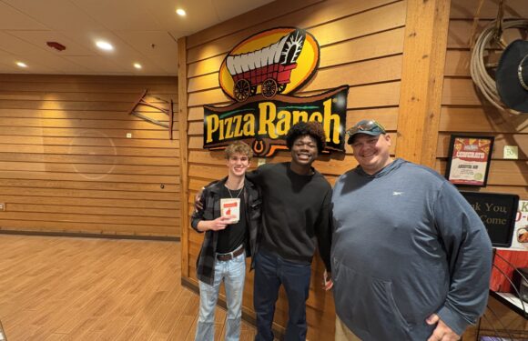 Three smiling young men stand together inside a pizza restaurant with a Pizza Ranch sign on a wooden wall behind them.