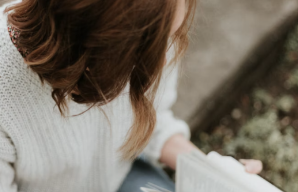 A woman with wavy, shoulder-length hair, wearing a white sweater, sits outdoors holding an open book. The background includes a blurred path and greenery.