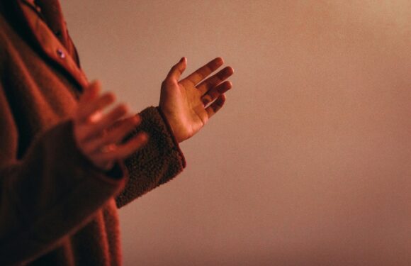 Part of a person’s arms and hands are visible, clapping or gesturing with palms facing outward, wearing a brown, textured jacket. The background is plain.