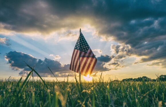 American flag waving in grassy field at sunset with dramatic clouds and sun rays in the background