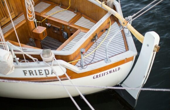 Vintage wooden sailboat docked on dark water, showing name “FRIEDA” and “GREIFSWALD” on the hull with rigging and railings visible.