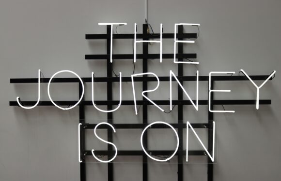 A neon sign on a gray wall displaying the words "THE JOURNEY IS ON" with a black grid structure behind the sign.