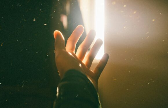 A hand reaches towards a bright, vertical light beam with particles floating around in the air, creating a dramatic and luminous effect.