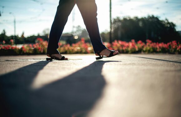 Person walking on a sidewalk wearing open-toe heeled sandals, with flowers and trees in the background and sunlight creating a long shadow.